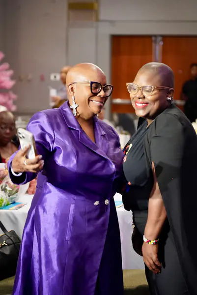 Two bald people pose for a selfie with one person wearing a purple jacket and the other person wearing a black top.