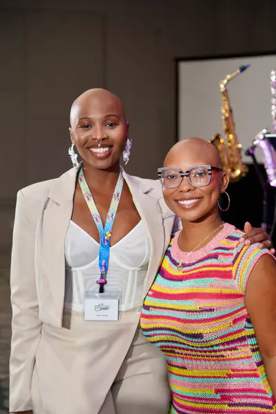 Two bald people stand together smiling for a photo while wearing a beige blazer over a white top with a conference lanyard and a colorful striped knit top.