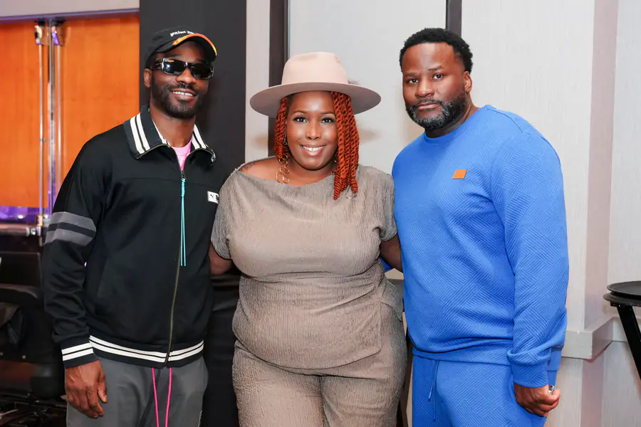 Three people stand together for a photo as a person in a black jacket and cap, a person in a beige hat and matching outfit, and a bald person in a blue sweater pose for the camera.