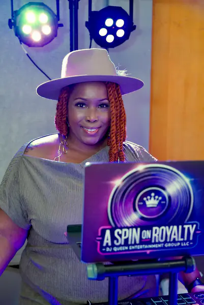 A bald person wearing a wide-brimmed hat and textured braids operates a DJ controller behind a laptop featuring a sticker that reads A Spin on Royalty DJ Queen Entertainment Group LLC.