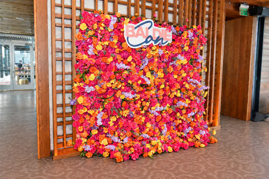 A wall of pink, orange, and yellow flowers serves as a backdrop for a sign at the top that reads Baldie Con.