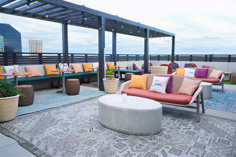 People gather on a rooftop lounge terrace where they sit on outdoor sofas and at tables decorated with pillows featuring the bald branded text.