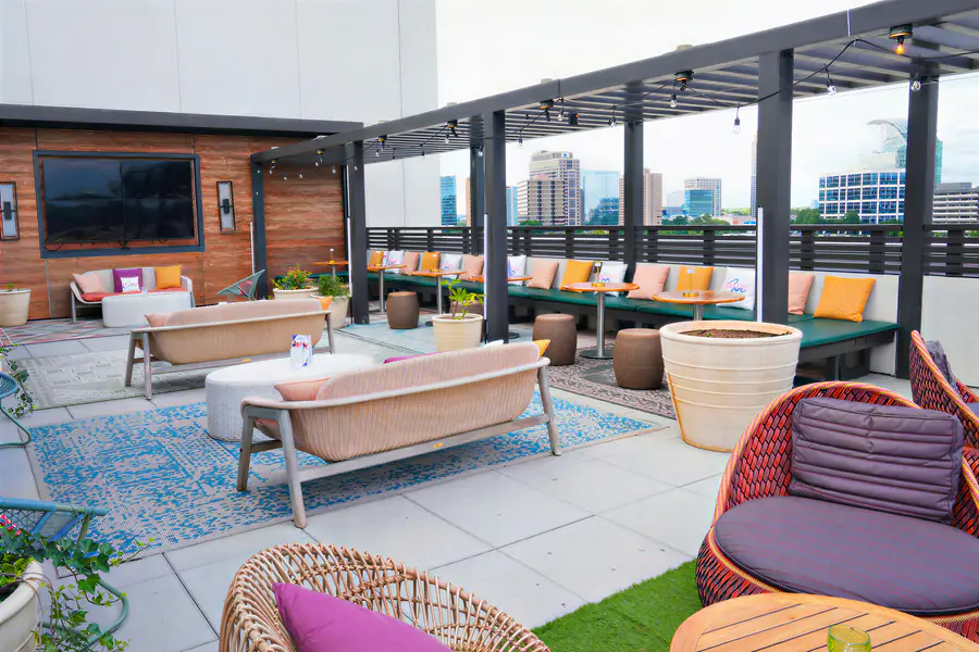 People who are bald gather in an outdoor rooftop lounge with modern seating, patterned rugs, and potted plants while looking out over a city skyline.