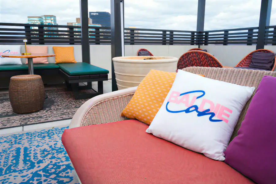 An outdoor lounge area on a rooftop features wicker furniture with cushions and a white pillow printed with the text Baldie Con.