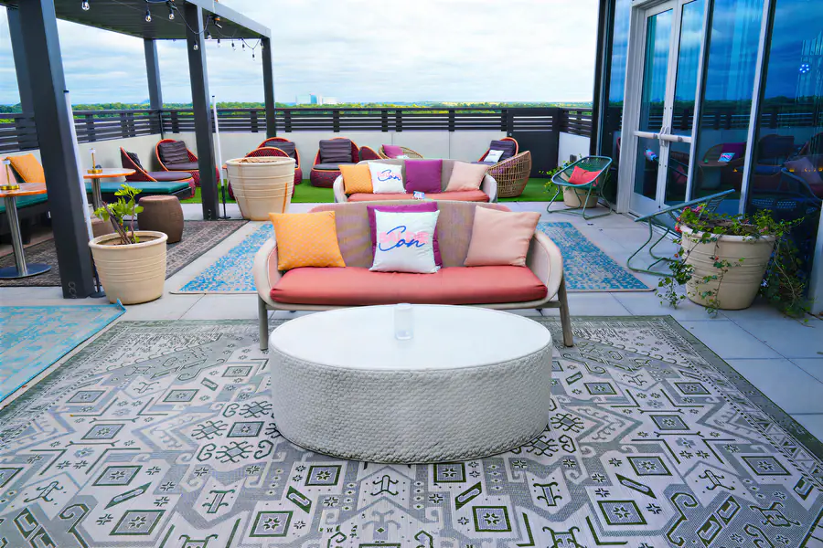 People who are bald gather in an outdoor rooftop lounge area featuring sofas and chairs with pillows, including two white pillows that each display the word Con in blue and pink script.