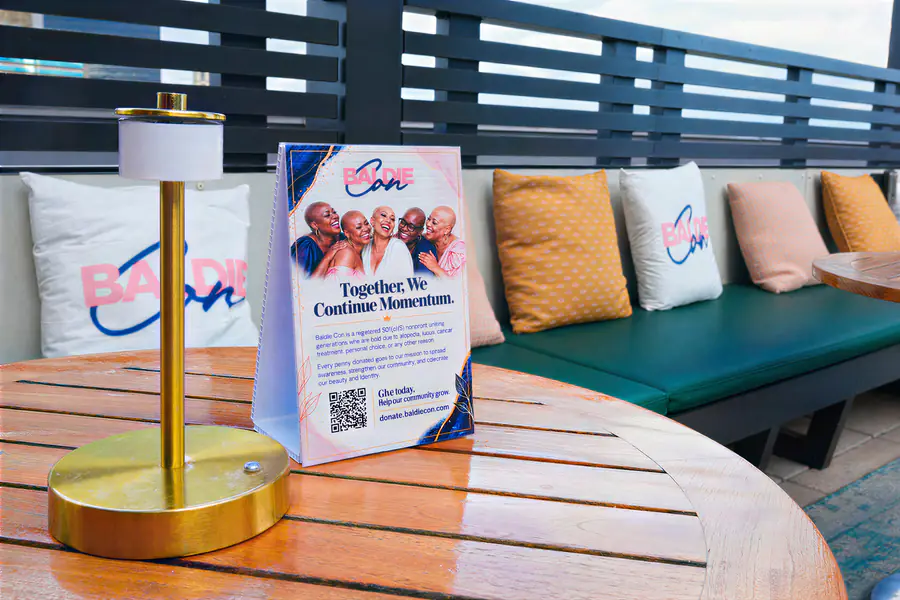 Five bald people smile together on a table tent promoting Baldie Con on a round table.