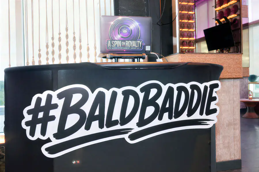 A person is operating a DJ booth that displays a large hashtag sign reading #BALDBADDIE and a laptop with a circular graphic featuring a crown and the text A SPIN ON ROYALTY at a conference for bald people.