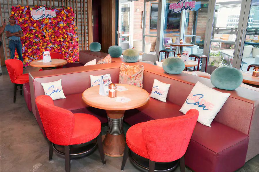 A bald person stands in front of a floral backdrop where they are surrounded by seating areas decorated with pillows bearing the Baldie Con logo.