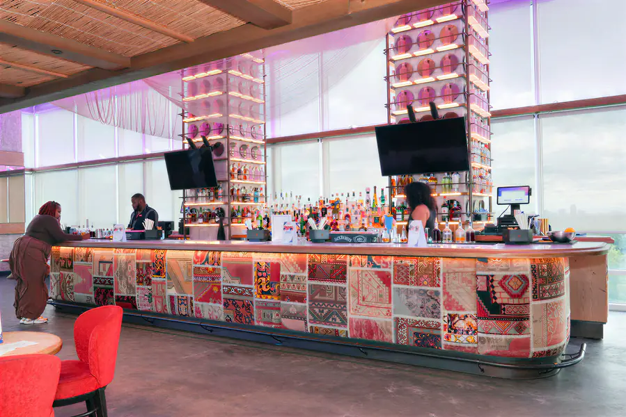 Three bald people stand behind a long bar with a patterned patchwork front in a brightly lit room as they work.
