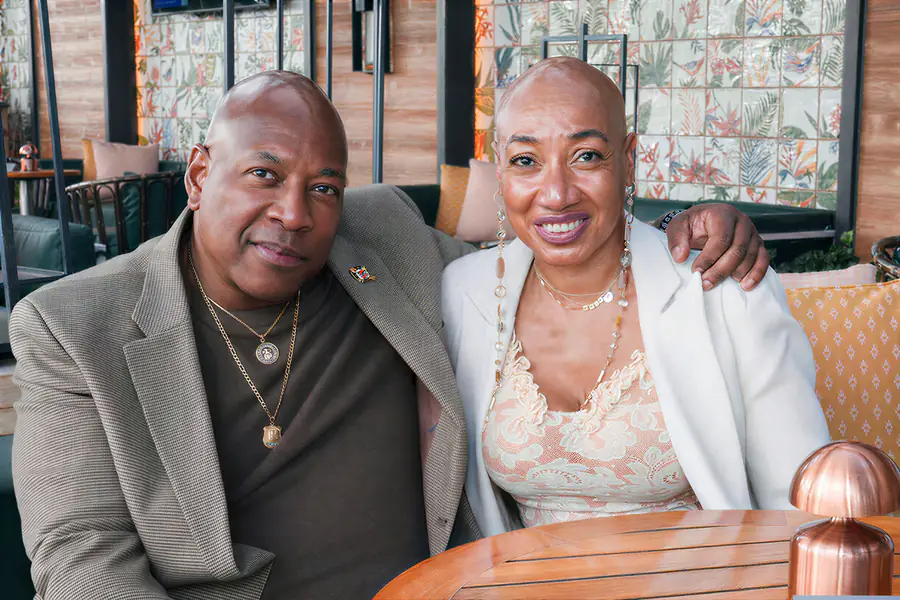 Two bald people sit together and smile at the camera while one person wears a tan blazer over a brown shirt and the other wears a white blazer over a lace top.