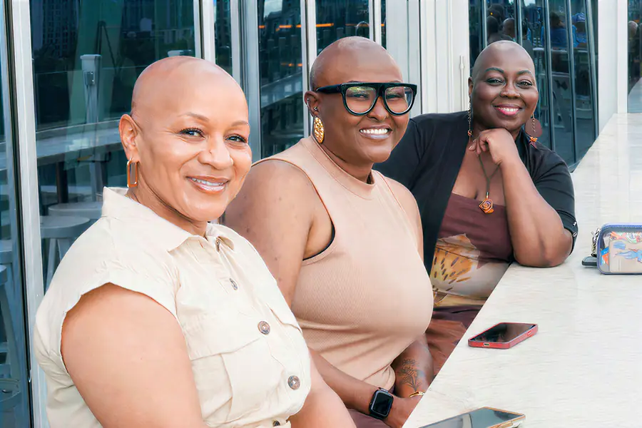 Three bald people smile while sitting together at a table.