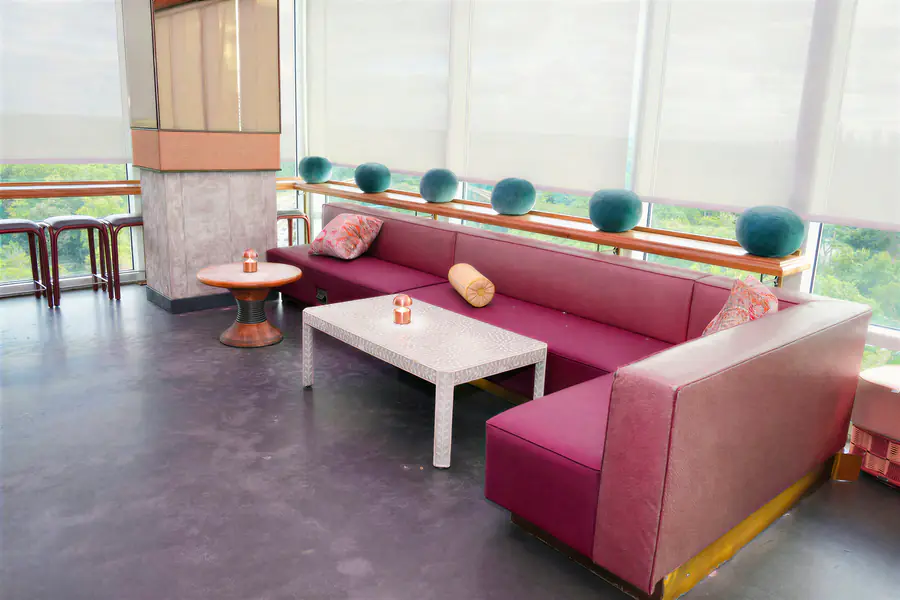 A large, L-shaped magenta sofa with round pillows sits in a bright lounge area next to a white table and a small round wooden table where a person who is bald rests.