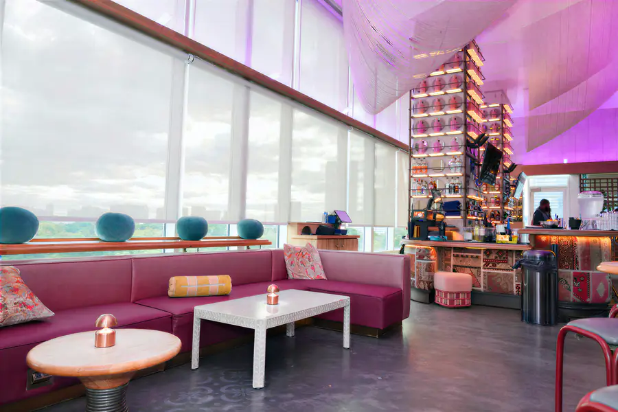 Bald people relax in a modern lounge area featuring a magenta sectional sofa, a white coffee table, and a tiered bar display with rows of small pink containers.