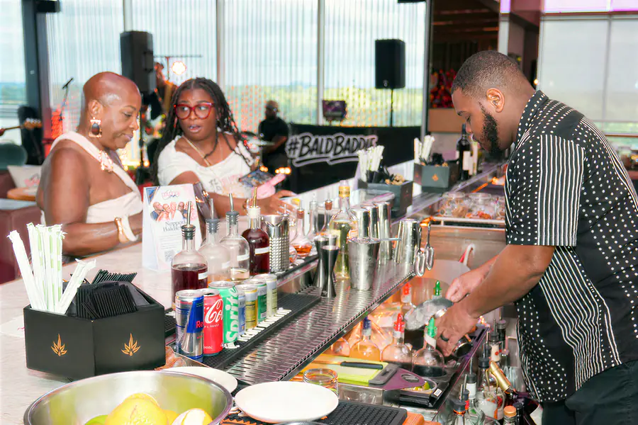 A bartender prepares drinks for two bald people sitting at a bar in front of a backdrop reading #BALDBADDIE.