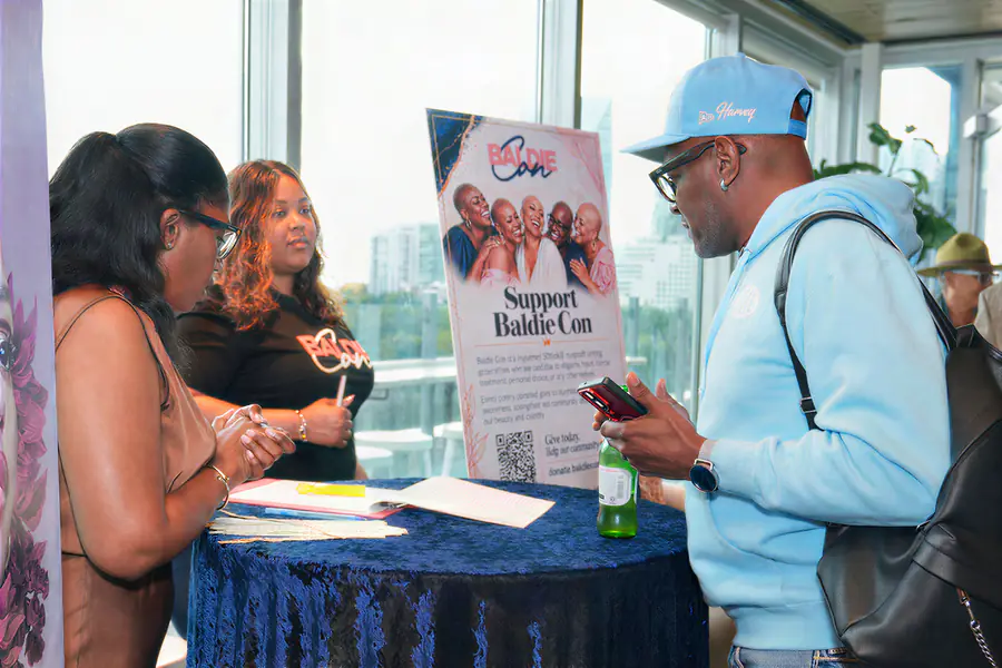 A person wearing a light blue hoodie and a baseball cap holds a phone and talks to two bald people who stand behind a table with a Baldie Con event sign.