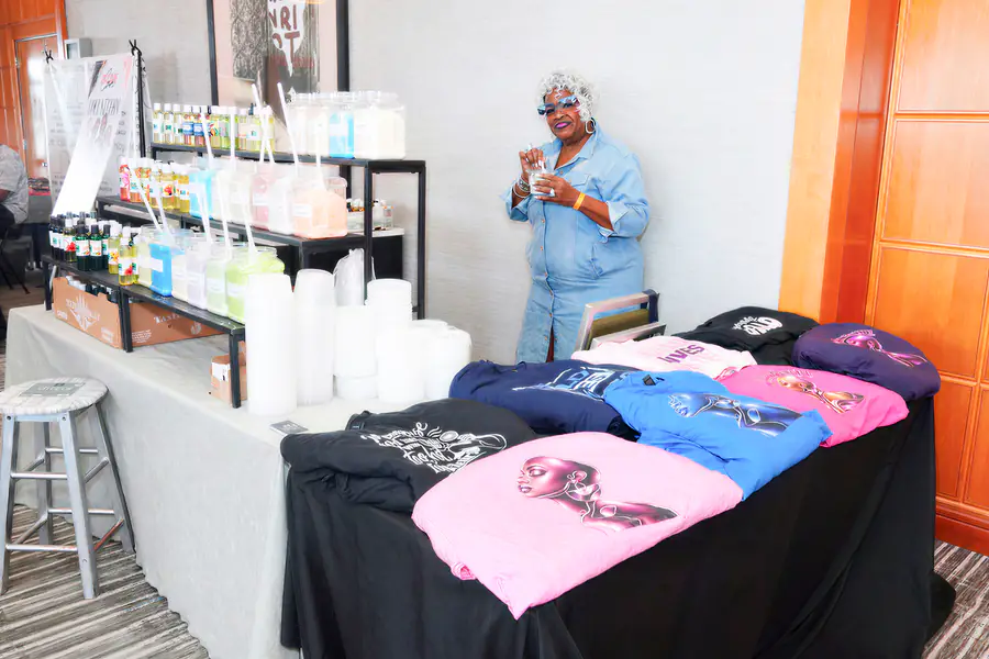 A bald person stands behind a table displaying colorful bottles and folded shirts with illustrations of bald people while they hold a small container.