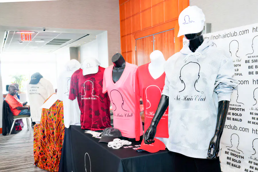 Mannequins are positioned at a conference table to display hoodies, t-shirts, and hats that feature the No Hair Club logo and the text be smooth be bald.
