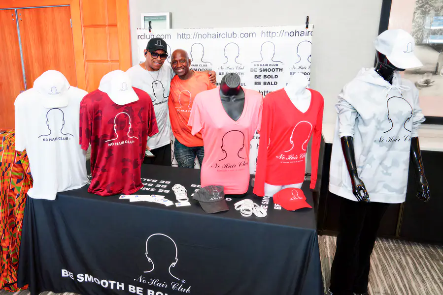 Two bald people stand behind a display table covered in apparel featuring the No Hair Club logo and text.
