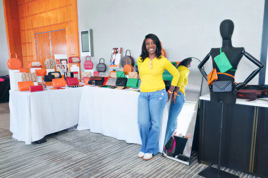 A bald person stands behind a table covered in various colorful handbags and clutches while wearing a yellow long-sleeved shirt and blue jeans next to a mannequin displaying small purses.
