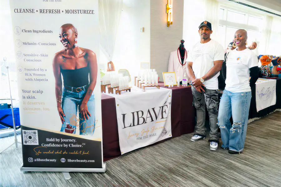 Two bald people stand beside a display table for Libavé scalp products while a large banner featuring a bald model hangs behind them.