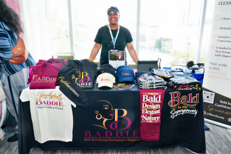A bald person stands behind a table covered with hoodies, t-shirts, and hats featuring the brand name B.A.D.D.I.E. and the slogan Bald Is My Superpower.
