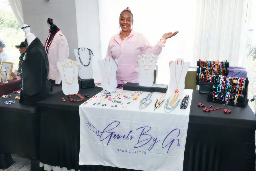 A bald person in a pink tracksuit gestures toward a display of necklaces, earrings, and bracelets on a table covered with a banner that reads Gewels By G Hand Crafted.