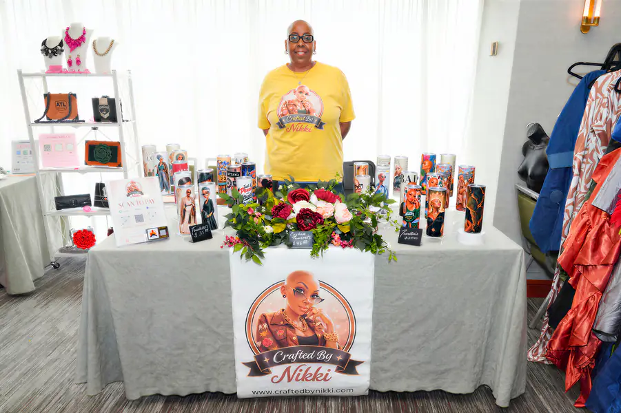 A bald person stands behind a table covered in decorative tumblers and a banner that reads Crafted By Nikki.