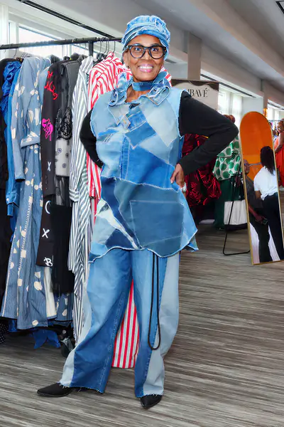 A smiling bald person stands with their hands on their hips in front of a clothing rack while wearing a patchwork denim vest, matching pants, and a denim head wrap.