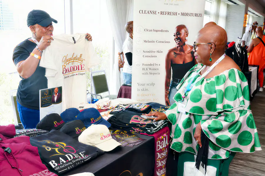 A person displays a white t-shirt reading Unapologetically Baddie to a bald person wearing a green and white polka dot top at a merchandise table featuring clothing branded with the word Baddie.