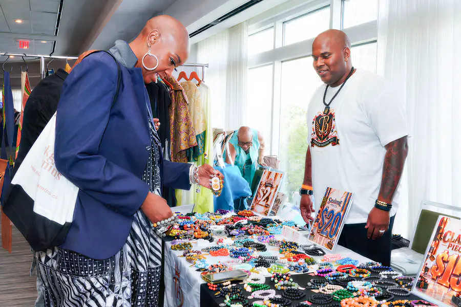 A bald person looks at a table of beaded bracelets while another bald person stands behind the table, and both people wear casual clothing.