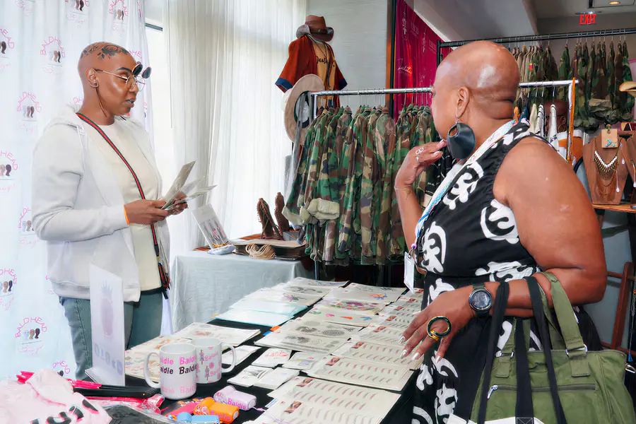 Two bald people stand at a market table with items on display including a mug with the word Baldie printed on it.