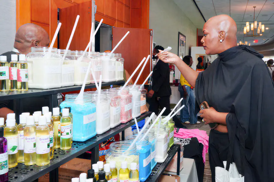 A bald person wearing a black top reaches toward a row of jars filled with colorful powders and scoops at a product display table.