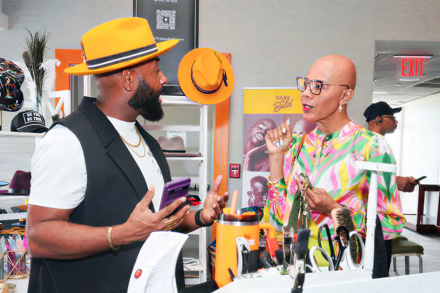 A person in a yellow hat and black vest speaks to a bald person wearing glasses and a colorful patterned shirt at a display table featuring a Dare to be Bald banner.