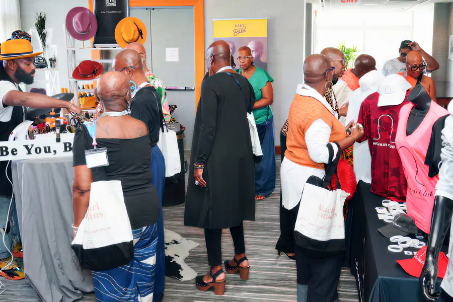 People who are bald walk between tables and browse displays of hats and apparel printed with the text No Hair Club.