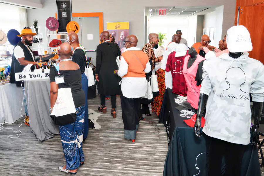 A group of bald people browse merchandise at tables featuring hats, shirts, and hoodies with text that reads No Hair Club.